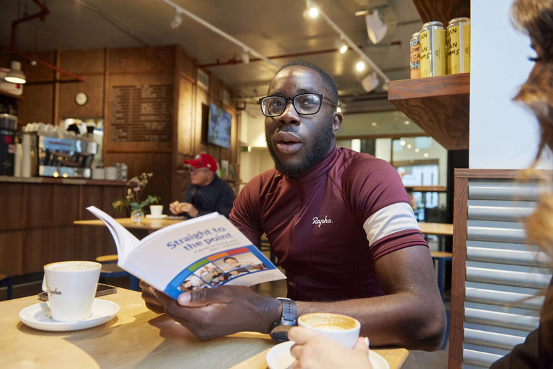 A man in a cafe, wearing a cycling top and reading a book produced by Breakthrough T1D UK about type 1 diabetes